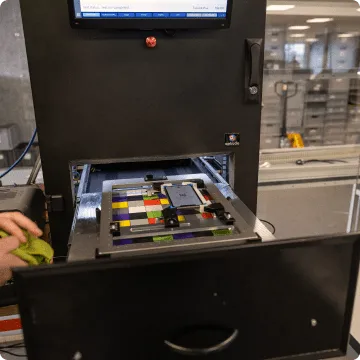 Ein gebrauchtes iPhone liegt eingeklemmt auf bunten Kacheln in einer viereckigen Vorrichtung. Daneben sind zwei Hände zu sehen, die einen Putzlappen halten.